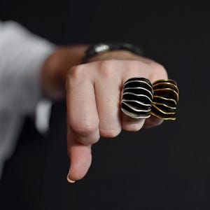 Big Bold silver and gold rings on a hand posing like a fist surrounded by black background with blurry white shirt
