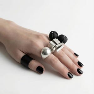 Hand wearing mixed black and silver statement rings: matte black sculptural sphere rings with small stud details, geometric silver block rings, a smooth silver dome ring, and a black vegan leather finger band; shown against a clean white background with black polished nails, minimal industrial aesthetic.
