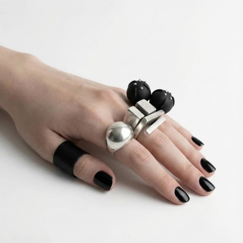 Hand wearing mixed black and silver statement rings: matte black sculptural sphere rings with small stud details, geometric silver block rings, a smooth silver dome ring, and a black vegan leather finger band; shown against a clean white background with black polished nails, minimal industrial aesthetic.
