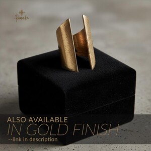 Gold statement ring with a geometric open-ended design displayed on a black velvet jewelry box, emphasizing its brushed metallic finish and sculptural form in soft natural light.