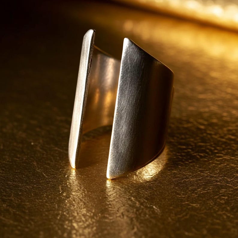 Close-up of a gold geometric open-ended statement ring placed on a reflective golden surface, highlighting its sculptural form and brushed metallic texture under warm light.