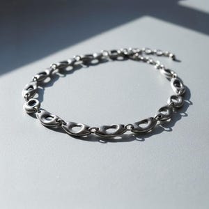 Silver sculptural chain necklace with oval open links displayed on a gray surface under natural sunlight. The smooth, organic forms cast soft shadows, highlighting its modern, architectural design — a unisex statement piece with minimalist industrial appeal.