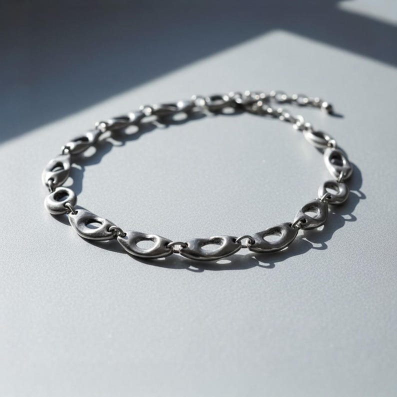 Silver sculptural chain necklace with oval open links displayed on a gray surface under natural sunlight. The smooth, organic forms cast soft shadows, highlighting its modern, architectural design — a unisex statement piece with minimalist industrial appeal.