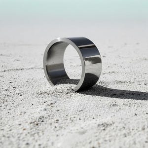 Polished stainless steel band ring standing on fine white sand under natural light. The smooth reflective surface and precise edges highlight its modern minimalist construction. Contemporary unisex jewelry design by Rannka with clean architectural lines.