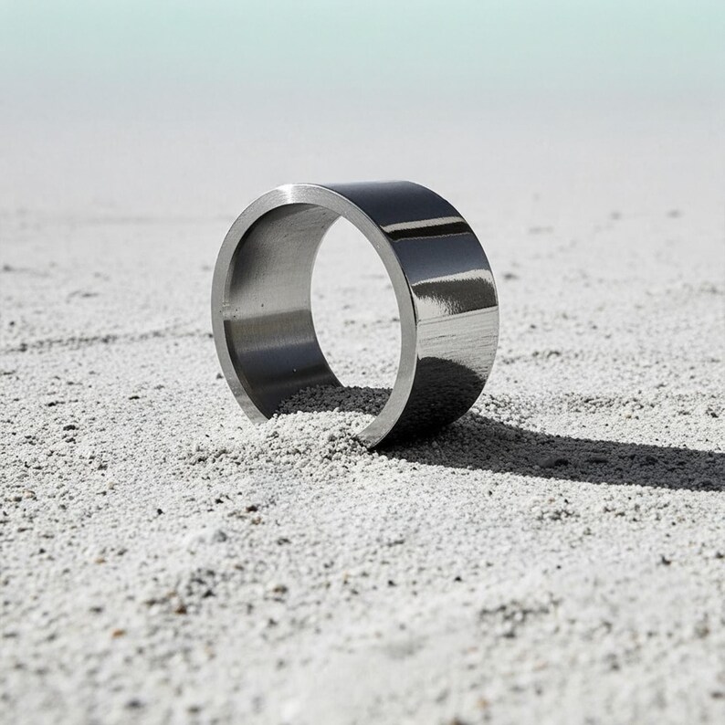Polished stainless steel band ring standing on fine white sand under natural light. The smooth reflective surface and precise edges highlight its modern minimalist construction. Contemporary unisex jewelry design by Rannka with clean architectural lines.