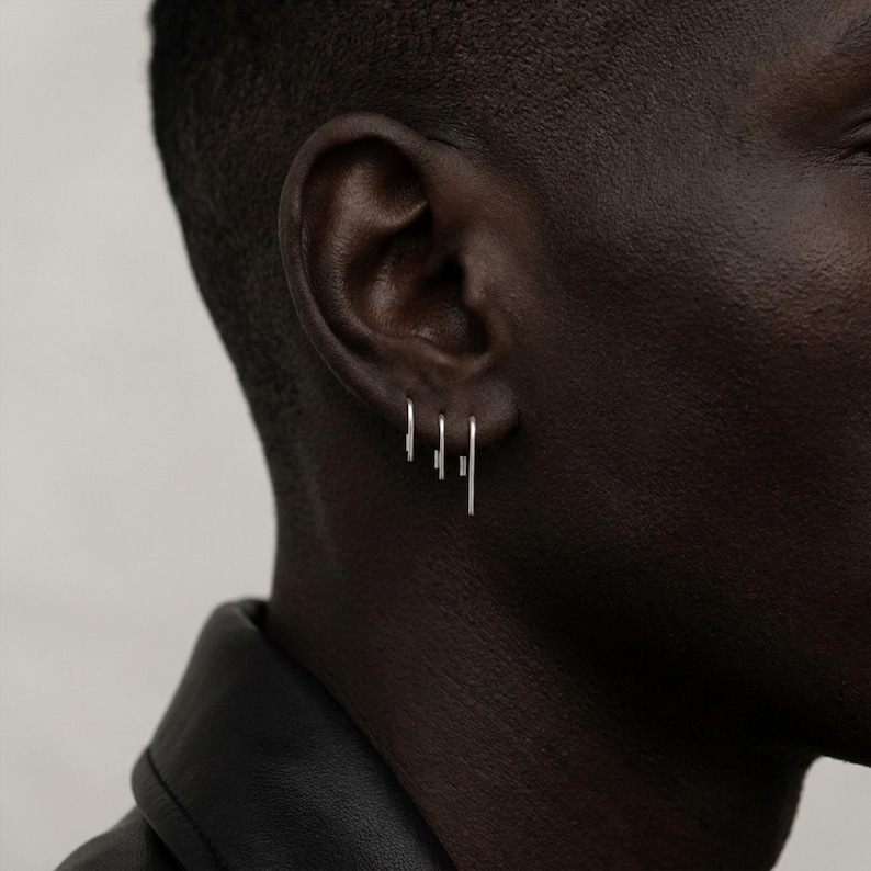 Minimalist sterling silver slip earrings on ear, small and medium bar drop earrings in stacked piercing styling, modern asymmetric ear stack, clean industrial jewelry look, subtle everyday silver earrings, close-up ear with multiple piercings, Rannka style, sleek and understated mood