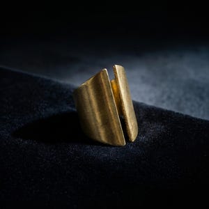 Modern sculptural gold ring shown in close-up as the focal piece, styled with a minimalist gold ear cuff and wide cuff bracelet, photographed on a dark textured surface with directional light highlighting the brushed metal finish and clean architectural form.