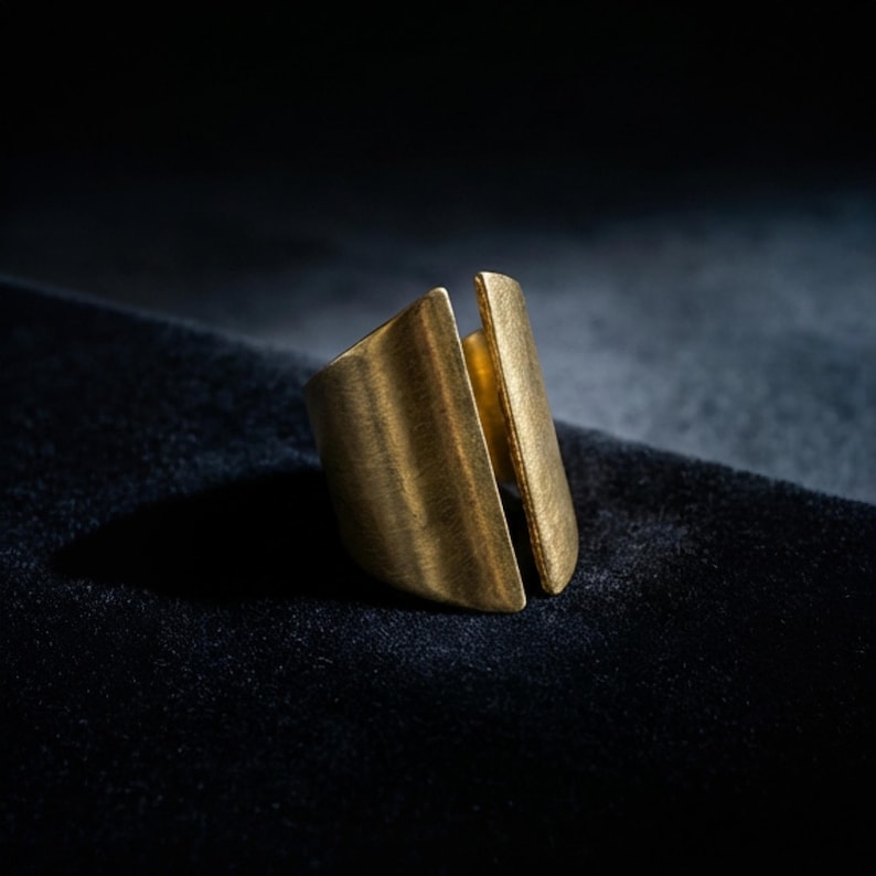 Modern sculptural gold ring shown in close-up as the focal piece, styled with a minimalist gold ear cuff and wide cuff bracelet, photographed on a dark textured surface with directional light highlighting the brushed metal finish and clean architectural form.