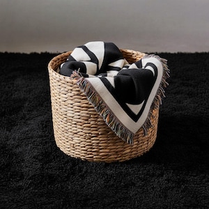 Folded black and cream geometric woven blanket with multicolor fringed edges placed in a natural wicker basket on a black textured rug. The tight weave and bold pattern create a modern, graphic contrast with the soft neutral tones of the space.
