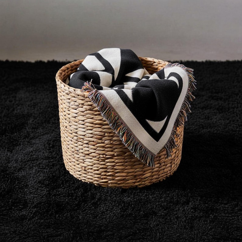 Folded black and cream geometric woven blanket with multicolor fringed edges placed in a natural wicker basket on a black textured rug. The tight weave and bold pattern create a modern, graphic contrast with the soft neutral tones of the space.