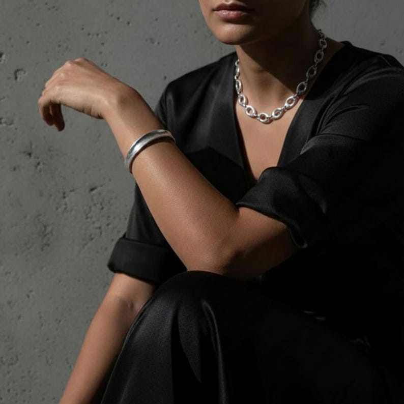 Editorial-style close-up of a model seated against a textured concrete wall, wearing a sculptural silver oval-link chain necklace and a smooth minimal silver cuff bracelet. Styled with a black tailored outfit, rolled sleeves, and soft directional light, highlighting clean lines, metal texture, and a restrained, architectural Rannka aesthetic.
