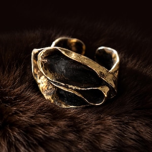 Könnte beinhalten: Ein offener Ring mit einer texturierten, goldfarbenen Metalloberfläche und einem dunklen, oxidierten schwarzen Innenbereich. Das Band hat eine unregelmäßige, organische Form mit gefalteten Kanten und einem verwitterten Finish.