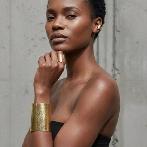 Portrait of a model wearing a wide brushed gold cuff bracelet, a sculptural open gold ring, and a minimalist gold ear cuff, hand resting under the chin, bare shoulders, matte metal jewelry with an architectural shape, modern editorial styling, Rannka jewelry.