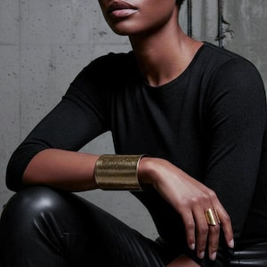 Editorial-style close-up of a model seated in black clothing, wearing a wide brushed gold cuff bracelet and an open sculptural gold ring, arm resting on bent knee, natural skin tones, matte metal texture, minimalist modern jewelry styling, architectural lines, contemporary luxury aesthetic, Rannka jewelry.