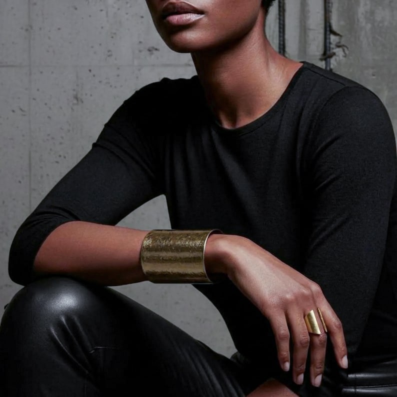 Editorial-style close-up of a model seated in black clothing, wearing a wide brushed gold cuff bracelet and an open sculptural gold ring, arm resting on bent knee, natural skin tones, matte metal texture, minimalist modern jewelry styling, architectural lines, contemporary luxury aesthetic, Rannka jewelry.
