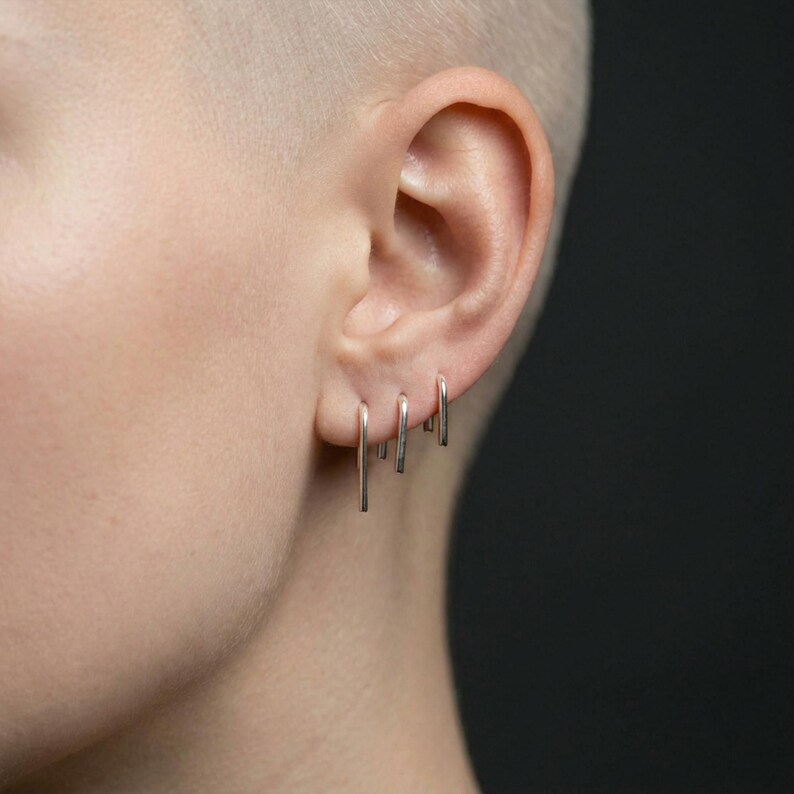 Sterling silver vertical bar earrings in mixed lengths, minimalist drop earrings worn in triple piercings, slim straight line earrings with clean edges, modern stacked ear styling, subtle asymmetric silver look, close-up ear detail on shaved head, sleek contemporary jewelry, Rannka aesthetic