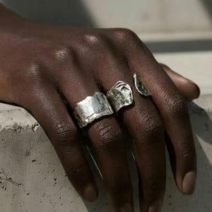 Puede incluir: Tres anillos de plata con diseños texturizados e irregulares se muestran en una mano. Los anillos tienen una apariencia cruda y martillada, uno de ellos con forma de lágrima. La joyería se presenta sobre un fondo neutro, resaltando el brillo metálico.
