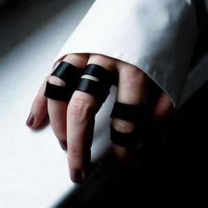 May include: Close-up of black leather finger rings. The rings are worn on multiple fingers, with a minimalist design. The rings are made of a matte black material. The background is a white surface and a white sleeve.
