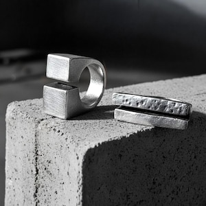 Two sculptural silver rings displayed on a textured concrete block. One features a bold open-square design, the other a split-bar hammered form. Both have a matte industrial finish, emphasizing Rannka’s modern, architectural jewelry aesthetic.