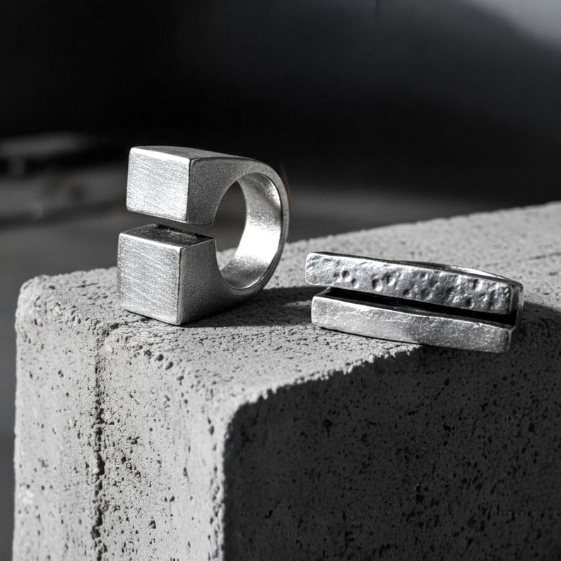 Two sculptural silver rings displayed on a textured concrete block. One features a bold open-square design, the other a split-bar hammered form. Both have a matte industrial finish, emphasizing Rannka’s modern, architectural jewelry aesthetic.