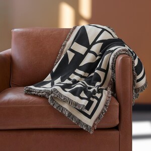 Black and cream geometric woven blanket with multicolor fringed edges draped over a brown leather armchair. The soft weave and bold pattern create a graphic contrast against the warm tones of the room, blending comfort with modern design.
