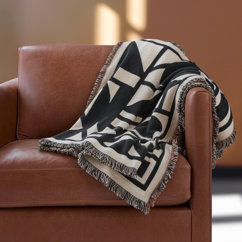 Black and cream geometric woven blanket with multicolor fringed edges draped over a brown leather armchair. The soft weave and bold pattern create a graphic contrast against the warm tones of the room, blending comfort with modern design.