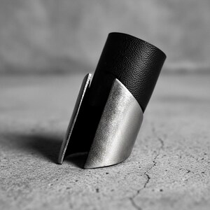 Close-up of a black vegan leather and steel statement ring placed on a concrete surface. The ring features a tall cylindrical leather band partially enclosed by matte steel plates with angled edges. Shot in monochrome tones with soft shadows and minimal industrial mood.