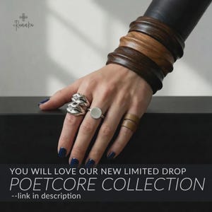 May include: A hand displays silver rings and wooden bracelets. The rings include a textured design and a flat circle. The bracelets are made of dark and light brown wood. The text reads "YOU WILL LOVE OUR NEW LIMITED DROP POETCORE COLLECTION --link in description."