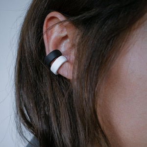 Black and white ear cuff duo worn on mid ear, no piercing ear jewelry hugging the ear edge, matte sculptural ear wraps with clean contrast, minimalist everyday ear styling shown in close-up profile, modern Rannka accessory look.