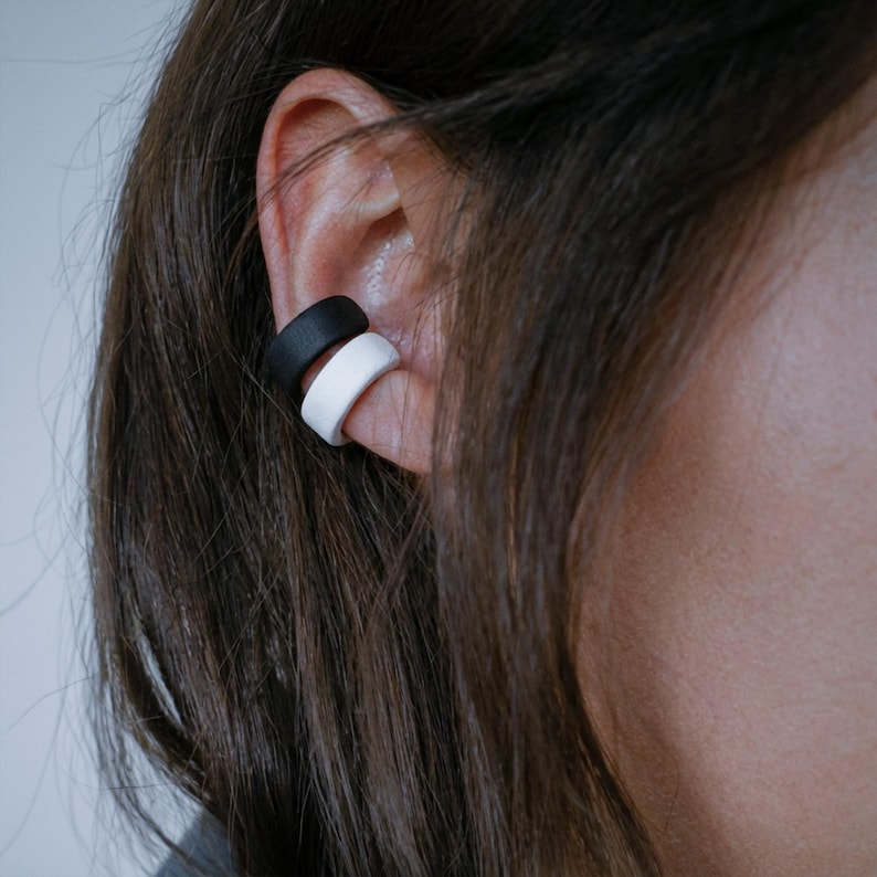 Black and white ear cuff duo worn on mid ear, no piercing ear jewelry hugging the ear edge, matte sculptural ear wraps with clean contrast, minimalist everyday ear styling shown in close-up profile, modern Rannka accessory look.