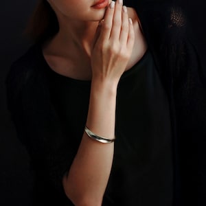 May include: A silver-toned cuff bracelet with a curved, organic design. The bracelet is worn on the arm of a person wearing a black top. The background is dark, emphasizing the jewelry's sleek, minimalist style.
