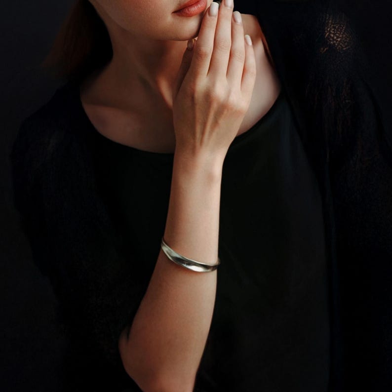 May include: A silver-toned cuff bracelet with a curved, organic design. The bracelet is worn on the arm of a person wearing a black top. The background is dark, emphasizing the jewelry's sleek, minimalist style.
