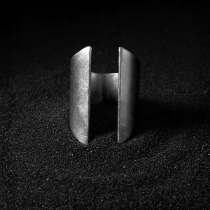 Close-up of a wide open-ended silver ring standing upright on a textured black surface, highlighting its matte metallic finish and minimalist sculptural design.