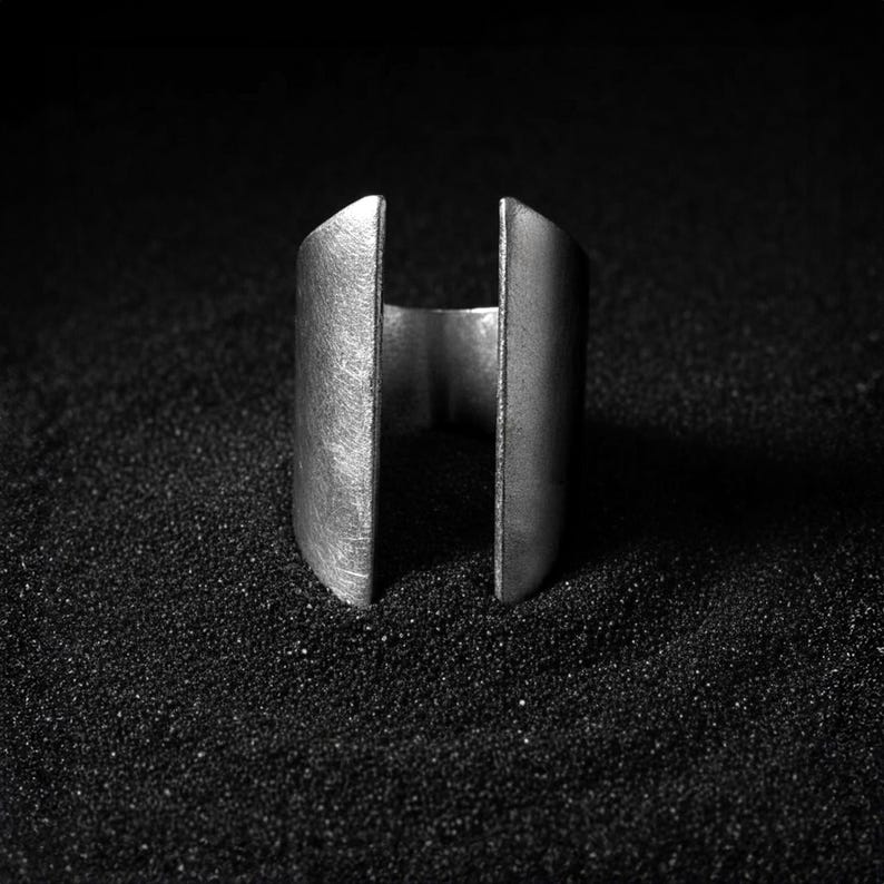 Close-up of a wide open-ended silver ring standing upright on a textured black surface, highlighting its matte metallic finish and minimalist sculptural design.