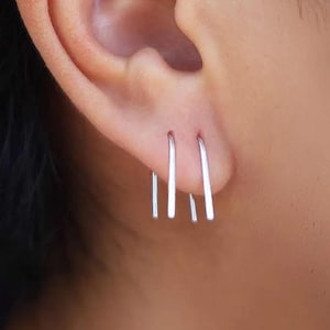 May include: Close-up of a person's ear with three silver bar earrings. The earrings are straight, thin, and have a minimalist design. The earrings are worn in the earlobe.