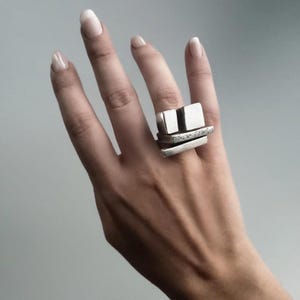 Brutalist silver ring worn on hand, featuring stacked geometric blocks with raw architectural form. Modern sculptural jewelry by Rannka, statement piece with bold rectangular layers. Minimal gray background highlighting the edgy and contemporary style of this gender-neutral accessory.