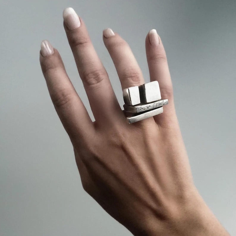 Brutalist silver ring worn on hand, featuring stacked geometric blocks with raw architectural form. Modern sculptural jewelry by Rannka, statement piece with bold rectangular layers. Minimal gray background highlighting the edgy and contemporary style of this gender-neutral accessory.
