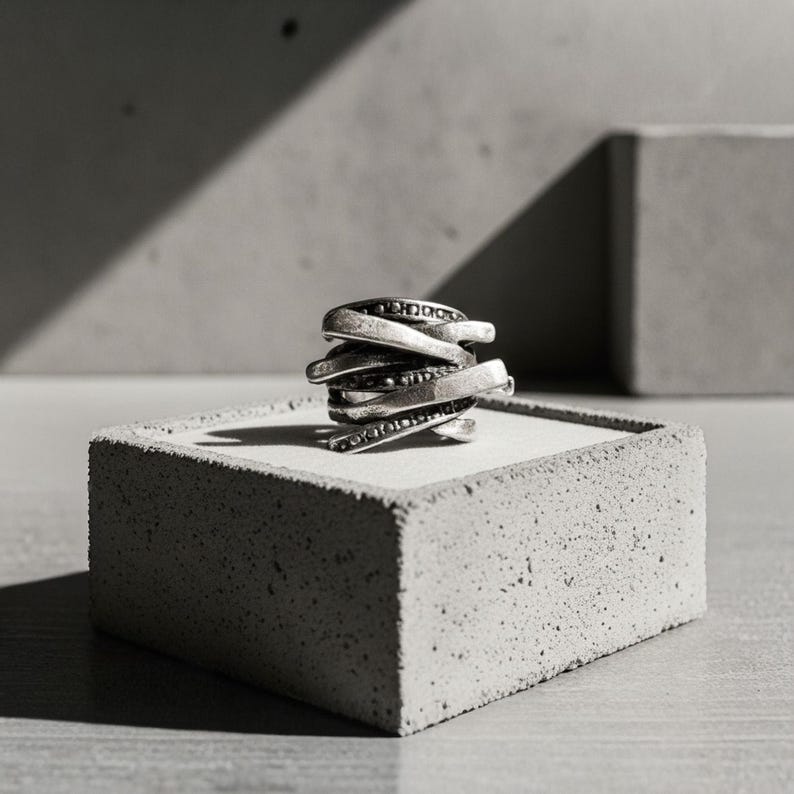 Sculptural oxidized silver ring with overlapping layered bands and dotted detailing, displayed on a concrete block under sharp directional light. The contrast of shadow and texture highlights the ring’s industrial and architectural form. Contemporary jewelry design by Rannka.