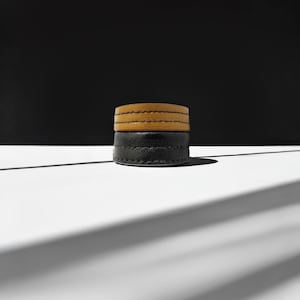 May include: Two stacked leather bracelets. The bottom bracelet is black, and the top bracelet is tan. Both bracelets have visible stitching and are circular in shape. The background is black and white, with a shadow cast across the white surface.