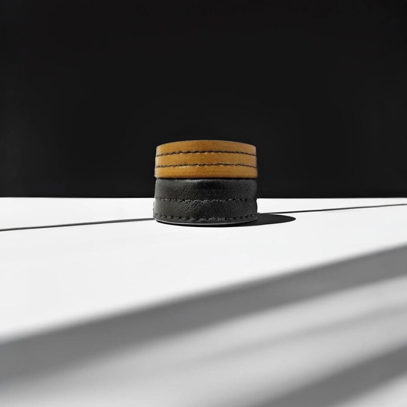 May include: Two stacked leather bracelets. The bottom bracelet is black, and the top bracelet is tan. Both bracelets have visible stitching and are circular in shape. The background is black and white, with a shadow cast across the white surface.