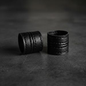 Two black vegan leather wrap rings placed side by side on a dark textured surface. Minimal leather ring cuffs with layered band construction and visible grain texture, photographed in low, moody lighting to emphasize depth and form. Sculptural contemporary jewelry with an architectural feel, shown off-hand to highlight material, thickness, and craftsmanship. Cruelty-free minimalist rings by Rannka, styled for modern stacking, everyday wear, and design-focused gifting.