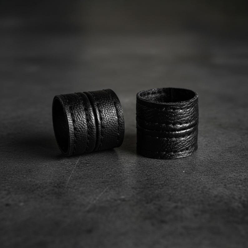 Two black vegan leather wrap rings placed side by side on a dark textured surface. Minimal leather ring cuffs with layered band construction and visible grain texture, photographed in low, moody lighting to emphasize depth and form. Sculptural contemporary jewelry with an architectural feel, shown off-hand to highlight material, thickness, and craftsmanship. Cruelty-free minimalist rings by Rannka, styled for modern stacking, everyday wear, and design-focused gifting.