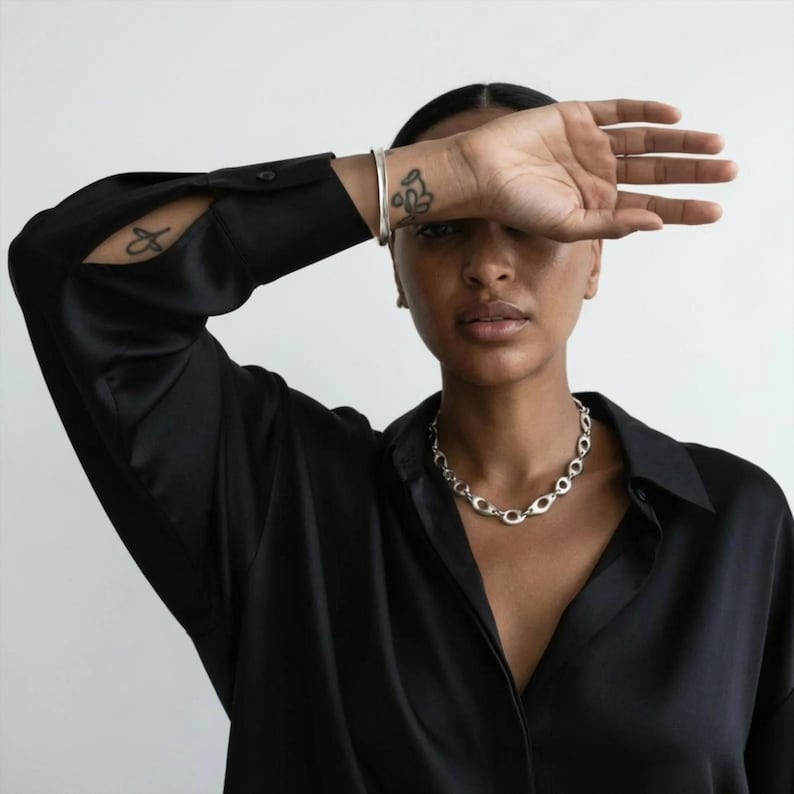 Portrait of a model in a black button-down shirt wearing a sculptural silver oval-link chain necklace and a minimal silver cuff bracelet. One arm is raised across the forehead, showing small wrist tattoos. Clean white background, soft natural light, modern editorial styling with a restrained, architectural feel aligned with Rannka’s aesthetic.