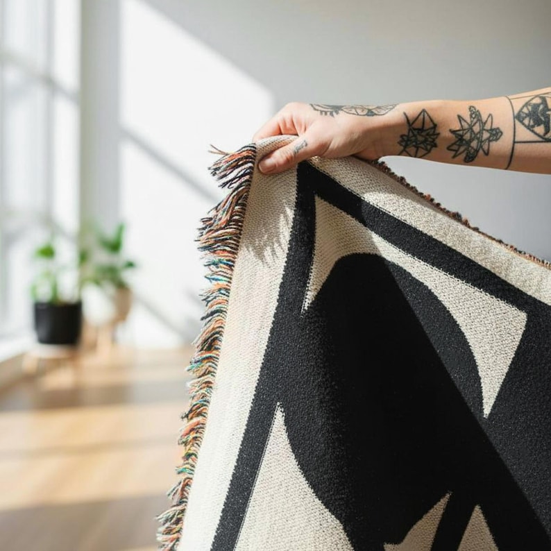 Close-up of a hand holding the corner of a black and cream geometric woven blanket with multicolor fringed edges. Sunlight highlights the textured cotton weave and bold abstract pattern, creating a warm and minimal interior feel.
