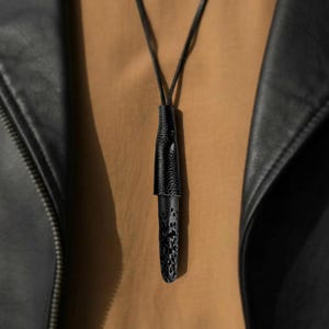 May include: A black pendant necklace with a leather cord. The pendant has a textured leather top and a patterned, cylindrical bottom. The necklace is displayed against a neutral background, with a black leather jacket visible on the sides.