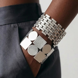 May include: Two silver bracelets are worn on a wrist. One bracelet has a woven brick pattern, and the other has a geometric design with circles and squares. The bracelets are made of a shiny metal.