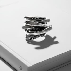 Close-up of a sculptural silver ring photographed on a white book under soft directional light. The ring features layered, blade-like bands with textured surfaces and dotted detailing, creating an organic, architectural look. Minimalist composition with sharp shadows and matte finish, matching Rannka’s industrial modern aesthetic.
