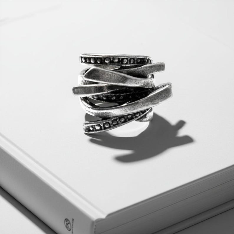 Close-up of a sculptural silver ring photographed on a white book under soft directional light. The ring features layered, blade-like bands with textured surfaces and dotted detailing, creating an organic, architectural look. Minimalist composition with sharp shadows and matte finish, matching Rannka’s industrial modern aesthetic.