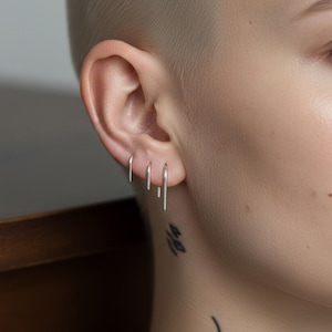 Sterling silver slim arc earrings in mixed lengths, delicate curved wire earrings worn across multiple piercings, lightweight minimalist drop wires, clean contour ear styling, modern understated silver jewelry, close-up ear with fine line tattoo detail, refined everyday look, Rannka aesthetic