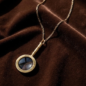 Può includere: Una collana con pendente color oro caratterizzata da un ciondolo a forma di lente d'ingrandimento circolare con manico cilindrico, sospeso a una sottile catenina color oro a maglia figaro. Il pendente ha una finitura metallica lucida.
